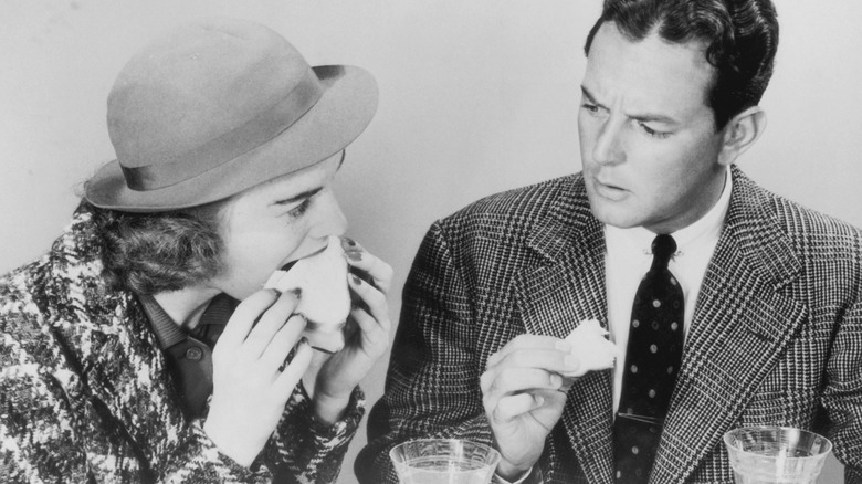 A black and white photograph of a man and a woman, dressed in the 1950s style, both eating sandwiches.