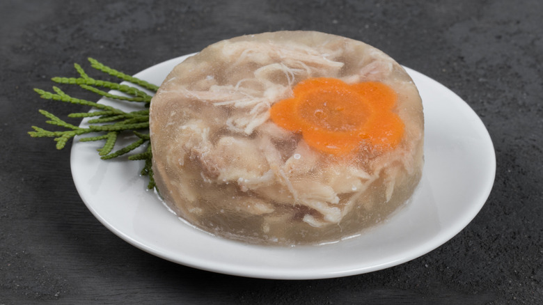 Some molded dish of jellied chicken and carrot, decorated with green herbs, sitting on a white plate on a grey surface.