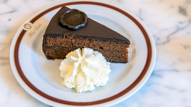 A slice of Sachertorte on a white plate next to fresh whipped cream