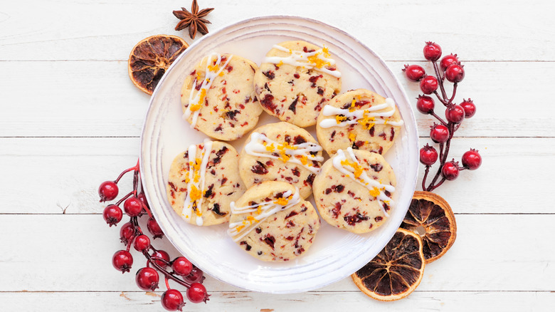 A white wooden table topped with a white plate featuring seven orange and cranberry-flavored with dried cranberries, dried oranges, and star anise scattered around