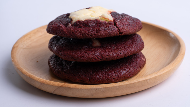 A light brown wooden plate with slightly raised edges is topped with three red-velvet cookies with a cream cheese filling