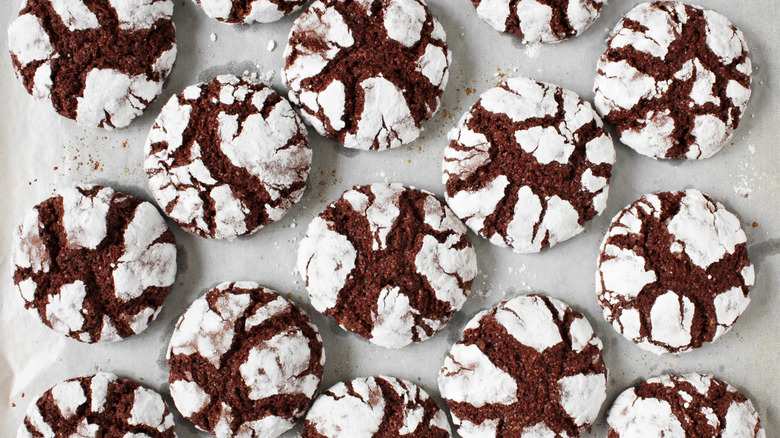 A pan with parchment paper is filled with freshly baked chocolate crinkle cookies topped with powdered sugar that has shifted during the baking process