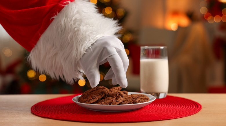 Santa Clause's arm and hand are seen grabbing a cookie from a white plate on top of a red placemat next to a single glass of milk. Holiday lights are twinkling in the background
