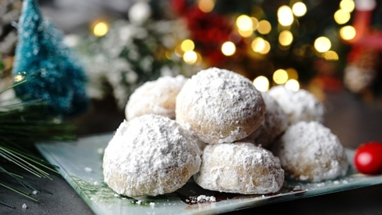 Pillowy lemon-flavored cookies coated in powdered sugar are stacked on top of one another on a plate in front of a background that is fuzzy but shows holiday lights