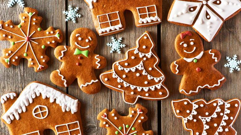 Gingerbread cookies in the shape of houses, snowflakes, trees, and people are decorated and scattered across a wooden table