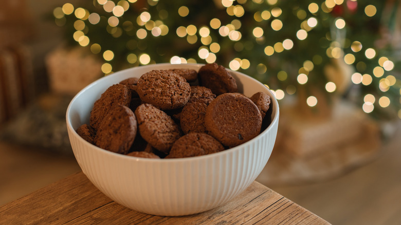 A white porcelain bowl is filled with mini oatmeal cookies and set on a table in front of a glowing Christmas tree