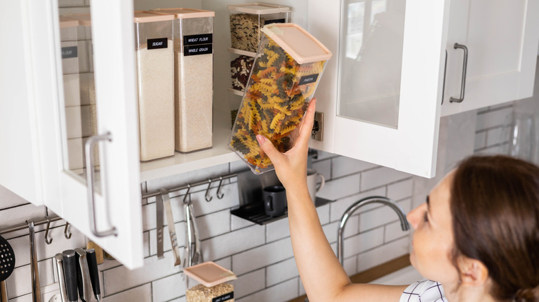 A woman puts a container of pasta into a cabinet.