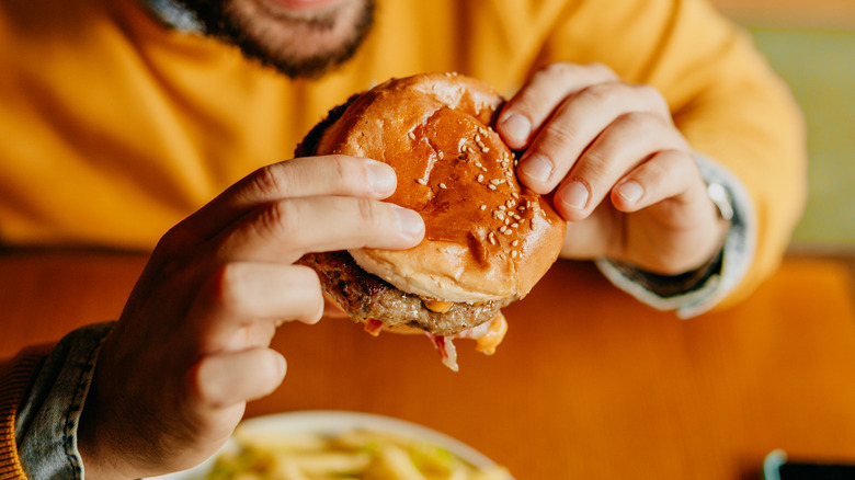 Bearded man in a yellow shirt holding a hamburger above a plate of french fries