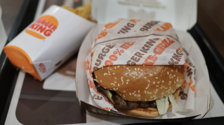 Burger King burger halfway wrapped in paper sitting in a cardboard carton on a tray beside a carton of french fries