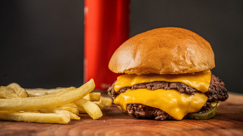 a double cheeseburger next to some fries with a drink out of focus in the background