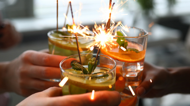 Three hands clinking cocktails garnished with lit sparklers