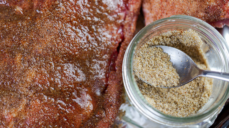 A spoon in a glass bowl of dry rub next to some rubbed meat
