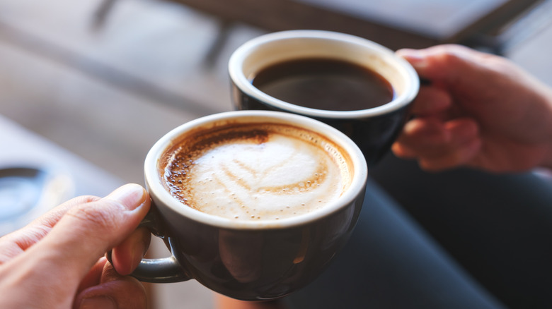 Two hands holding a mug of cappuccino and black coffee