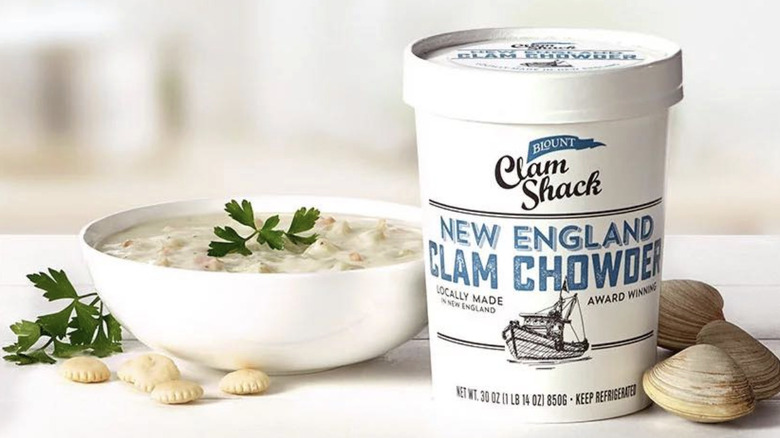 A cup of Blount Clam Shack New England Clam Chowder beside a bowl of the soup and some clams