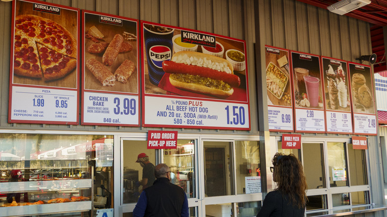 Customers waiting in line at the Costco food court with the classic menus displayed above the ordering windows