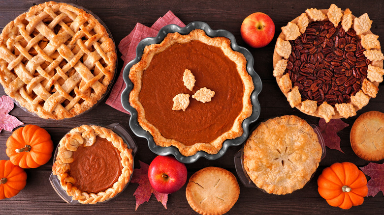 a variety of pie laid out on a wooden table with fruits and autumn leaves