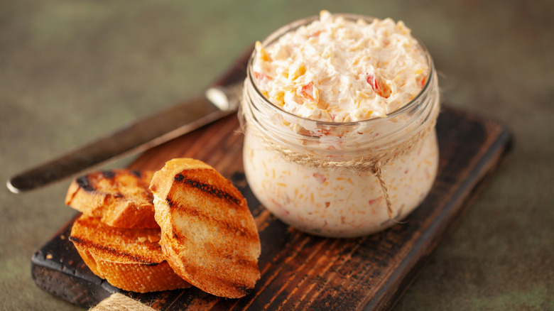 a jar of pimento cheese on a small cutting board with grilled bread and a knife