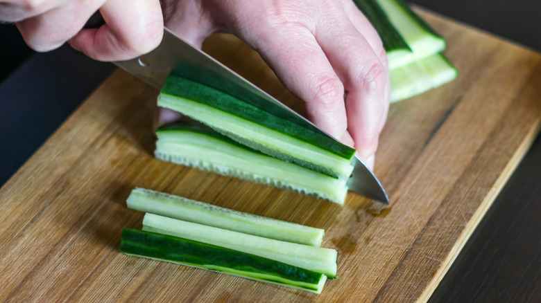 Person slicing cucumber sticks with santoku knife on wooden cutting board