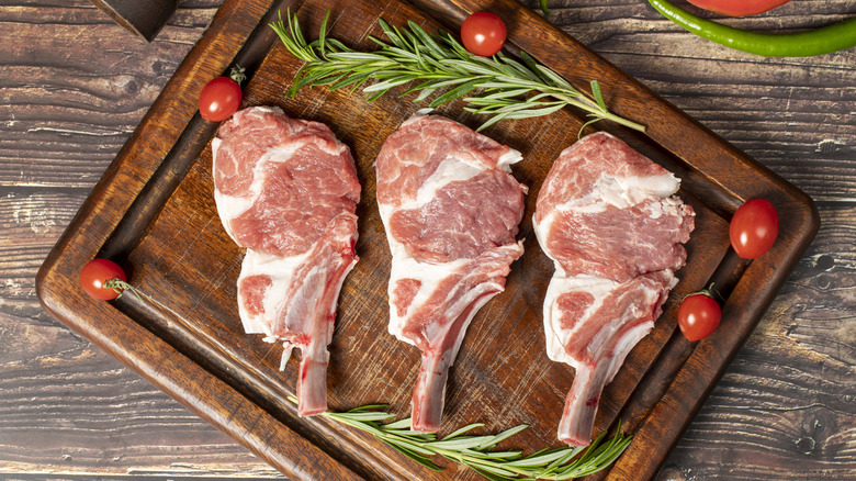 Raw lamb chops on a cutting board surrounded by rosemary and tomatoes