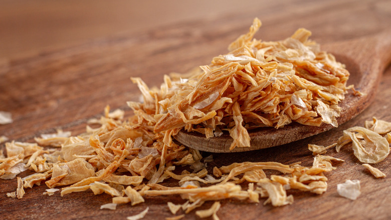 A wooden spoon full of crispy fried onion flakes that are spilling out onto a wooden table top around it