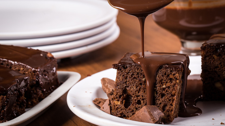 Chocolate sauce is being spooned over a piece of chocolate sheet cake on a plate.