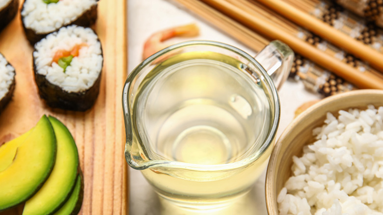 A measuring cup of vinegar sits between a bowl of cooked rice and prepared sushi rolls
