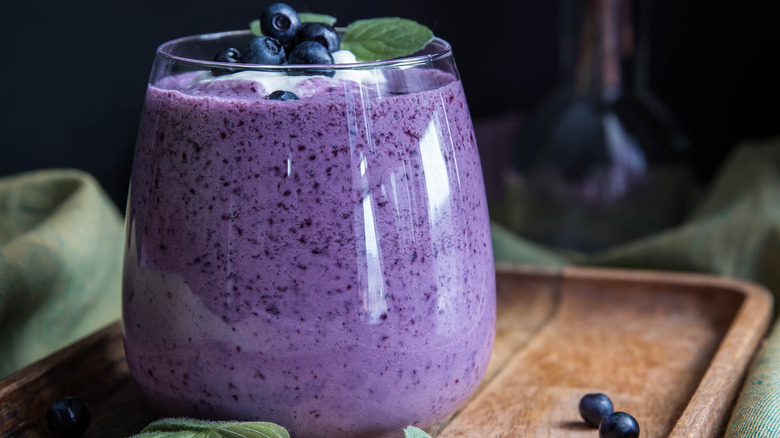 A blueberry smoothie in a clear glass on a wood tray