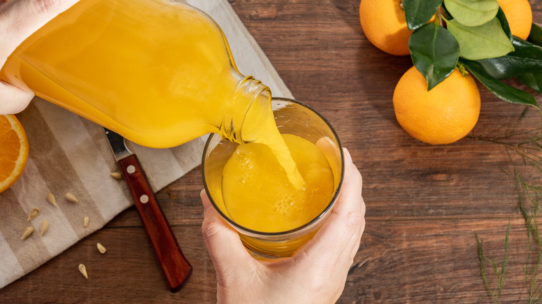 person pouring orange juice from a jug into a glass on a wooden table with whole oranges nearby