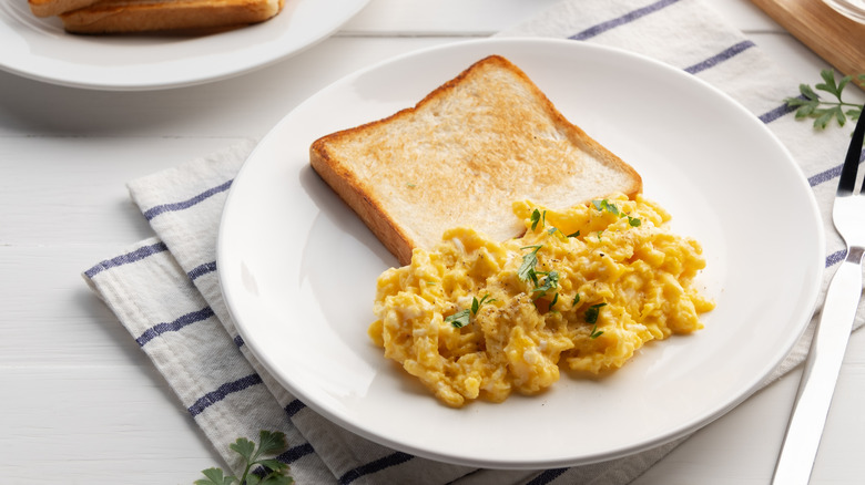 White plate of scrambled eggs with herbs next to slice of toast