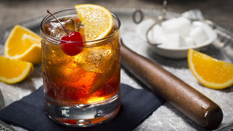 Closeup of a cocktail with a cherry and orange slices and sugar cubes behind