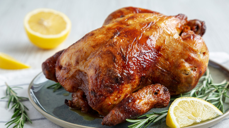 A golden roasted rotisserie chicken sitting on a tray with some lemon and rosemary