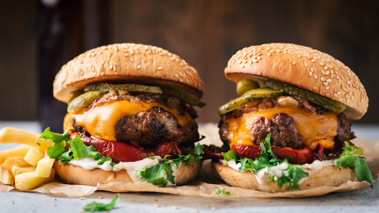 Two cheeseburgers with pickles, mayo, lettuce, and tomato next to french fries on gray surface