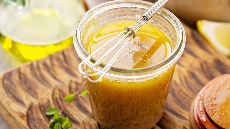 A glass jar full of a viscous-looking yellow marinade with a tiny whisk on top, sitting the top of wooden cutting board
