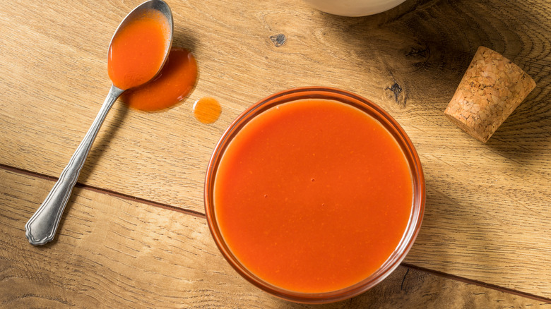 A bowl of Buffalo wing sauce on a wooden table next to a teaspoon of sauce and a cork