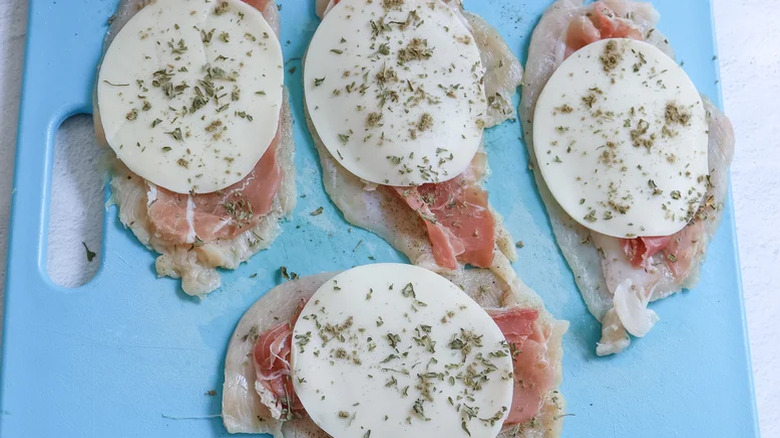 Four chicken breasts topped with prosciutto and provolone and herbs ready to be rolled for saltimbocca