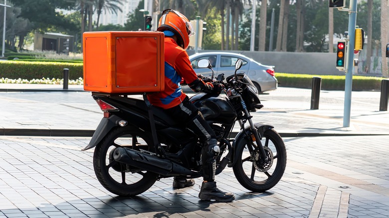 A deliver driver shown on a motorcycle with a large orange box behind them