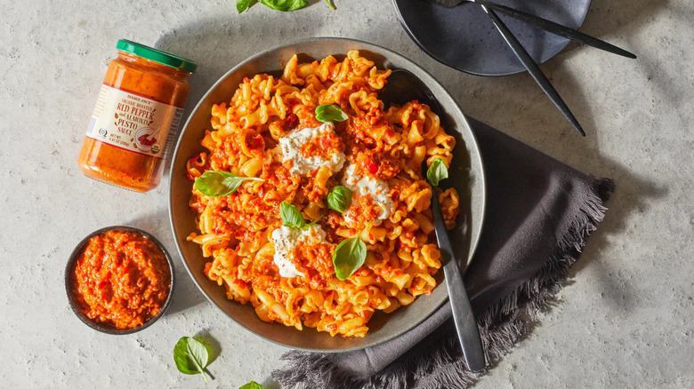 Jar and small bowl of Trader Joe's Roasted Red Pepper and Almond Pesto Sauce next to gray bowl full of red sauced pasta with cheese and basil leaves
