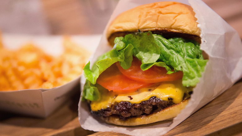 A cheeseburger with lettuce and tomato in a white paper bag next to a paper container of French fries