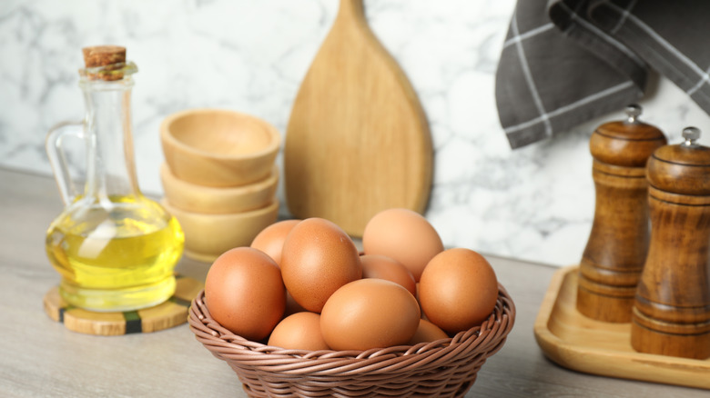 Eggs in wicker basket on counter, jug of olive oil, salt and pepper shakers, cutting board and wooden bowls around