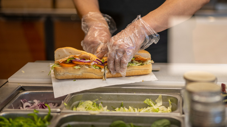 Pair of gloved hands cutting a submarine sandwich in half on the counter in front of bins of lettuce, onion, and green pepper