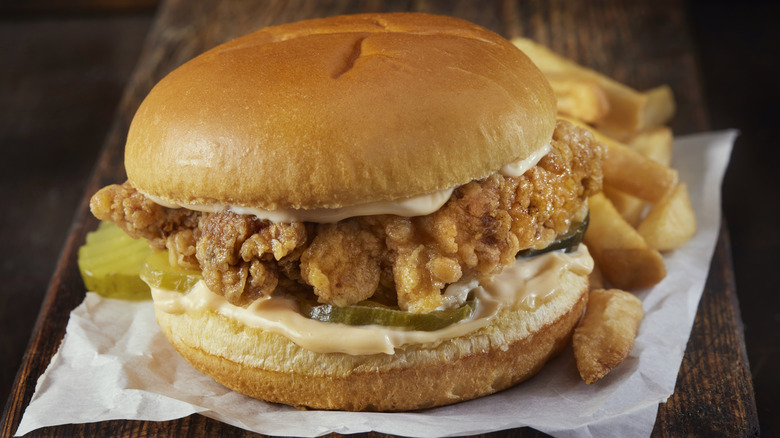 A crispy fried chicken sandwich on a bun with sauce and pickles, sitting on some paper with french fries