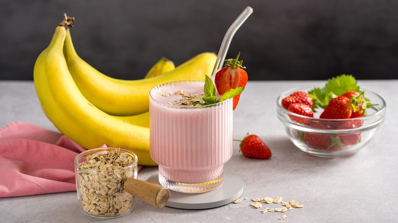 A pink smoothie in a glass surrounded by oats, bananas, and strawberries
