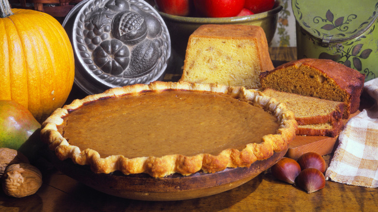A vintage photo of a pumpkin pie in front of pumpkins and breads