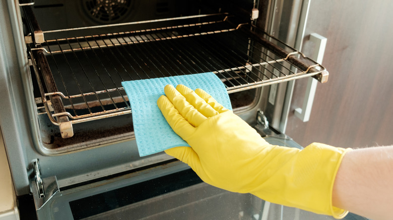A hand with a yellow rubber glove holding a blue cleaning sponge and wiping the rack of an open oven