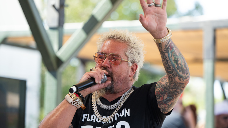 Guy Fieri speaking into a microphone with one hand up