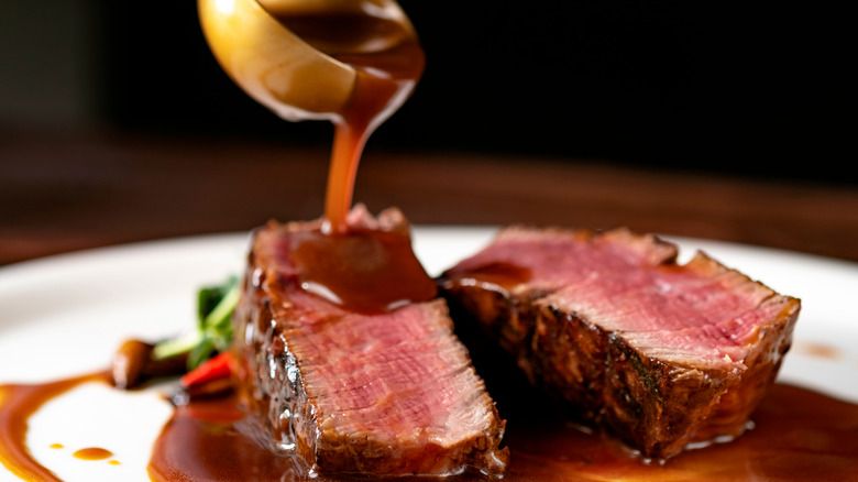 Ladle pouring brown demi glace sauce over sliced rare steak