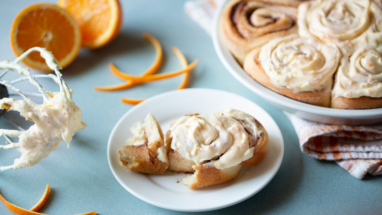 Orange cinnamon rolls with rich frosting on a table next to oranges and a whisk with cream cheese frosting on it