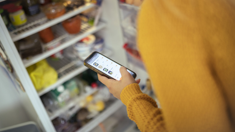 A person looks at a grocery shopping app on their phone with the refrigerator door open