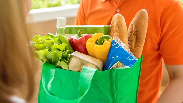Person in orange shirt delivering a bag of groceries to someone with long blonde hair