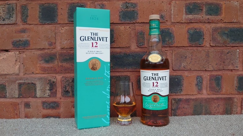 The box, a filled glass, and the bottle of The Glenlivet 12-year posed in front of a red brick wall